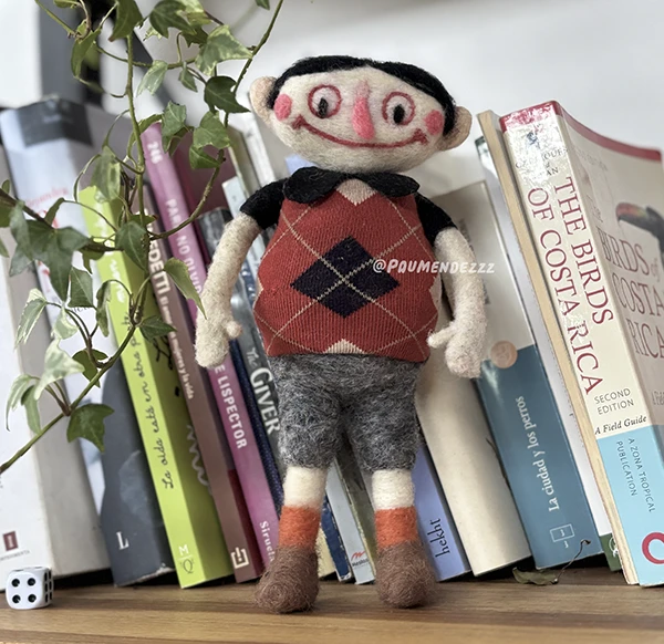 Muñeco en fieltro con chaleco rojo de rombos entre libros