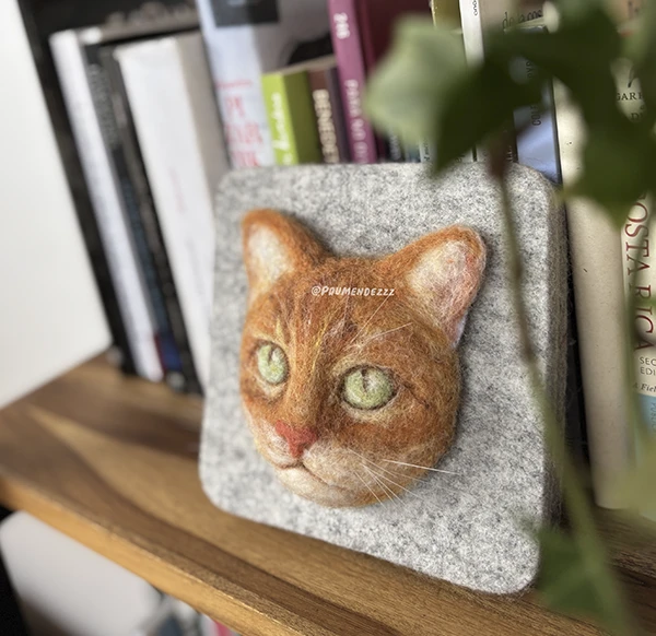 Cuadro en fieltro con la cara de un gato naranja apoyado en una repisa